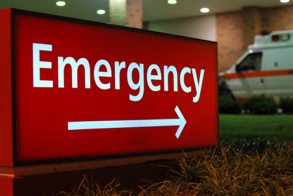 Red and white sign outside a hospital with the word "Emergency" pointing toward the emergency department; it's nighttime and an ambulance is parked outside 